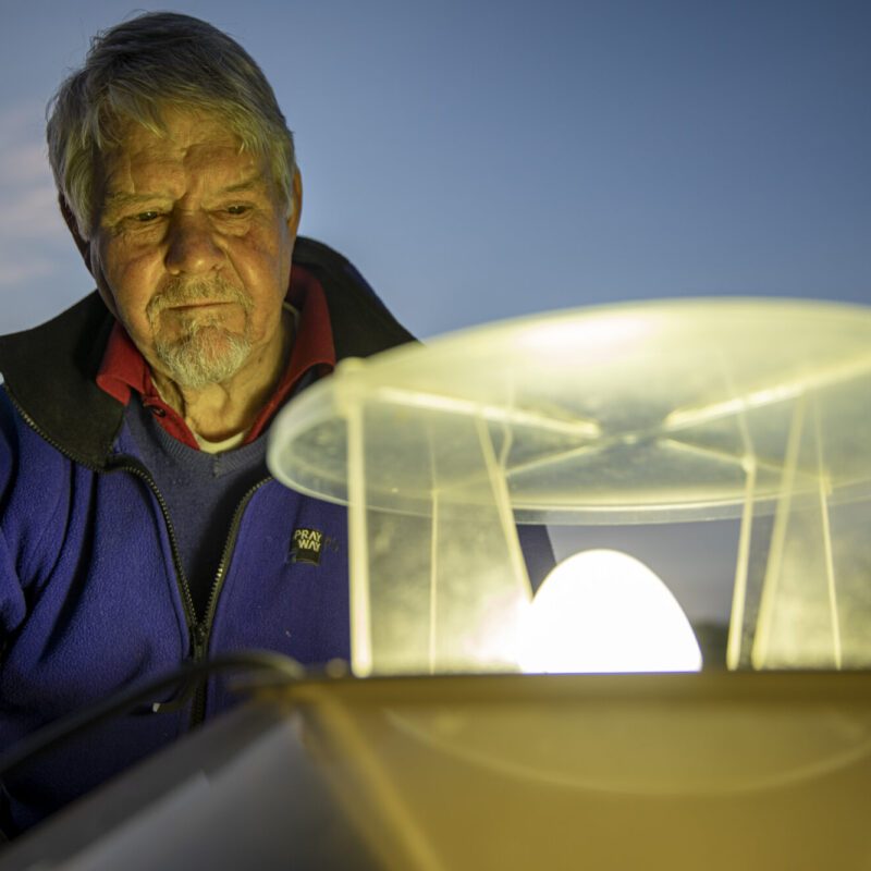 A man with short white hair and a goatee, in a blue fleece, observing a moth trap - a glowing light with a plastic lid above and a funnel below.