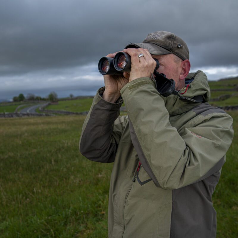 a man with stubble, wearing a green cap and rain coat, standing in a grassy field, looking through binoculars